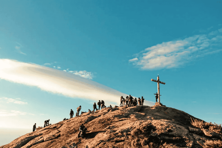 Grupo de pessoas no cume do Pico da Bandeira, próximo a uma cruz, em um dia de céu azul no Parque Nacional do Caparaó.
